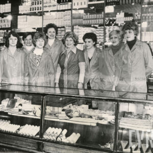 osiem kobiet w fartuszkach stoi za ladą sklepową i pozuje do zdjęcia, za nimi półki z artykułami kosmetycznymi