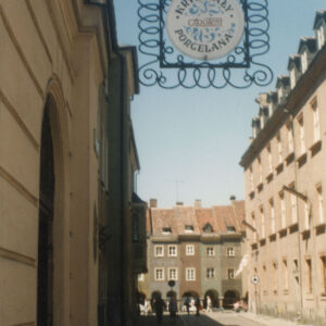 przy wejściu do budynku metalowy ozdobny szyld z nazwą sklepu, w głębi i z prawej kilkupiętrowe budynki