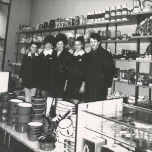 cztery kobiety i mężczyzna w ciemnych fartuchach stoją w przed półkami z artykułami gospodarstwa domowego, przed nimi na niskiej półce emaliowane garnki