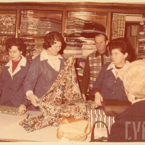 kilka osób stoi, wśród nich trzy kobiety w fartuchach z z jasnymi kołnierzykami przy ladzie, jedna z nich rozkłada belę tkaniny, za nimi drewniane regały z belami tkanin