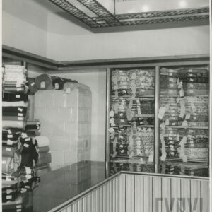 narożnik pomieszczenia, pod ścianami drewniane regały z belami tkanin, w narożniku kaflowy piec