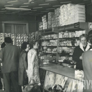 wnętrze sklepu, na regałach kartony z butami, przy ladzie sprzedawczyni w ciemnym fartuchu z jasnym kołnierzem i obsługiwani klienci