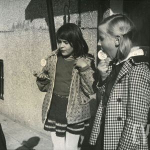 dwie dziewczynki noszące tornistry, jedzące lizaki na tle ściany bloku z wielkiej płyty