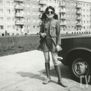 dziewczynka w sukience i okularach przeciwsłonecznych, stojąca przy samochodzie na tle bloku z wielkiej płyty