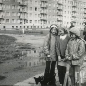 trzy dziewczynki, jedna z psem na smyczy, na tle bloku z wielkiej płyty