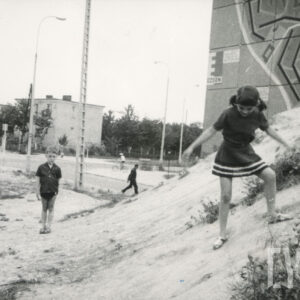 dzieci bawiące się na górce piasku na tle bloku z wielkiej płyty; w głębi skrzyżowanie ulic, dalej dom wolnostojący