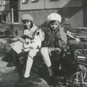 dwie dziewczynki w płaszczach z berecikami na głowie, jedna z nich trzyma lalkę, a obok stoi wózek dla lalek