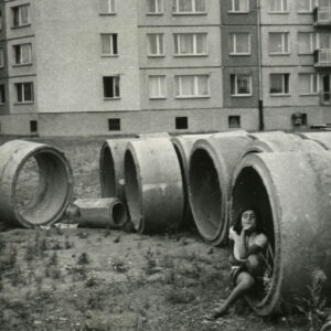 dziewczynka siedząca w betonowej obręczy na placu budowy, w głębi blok z wielkiej płyty