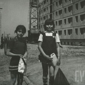 dwie dziewczynki stojące na tle budowanego bloku z wielkiej płyty, widoczne maszyny budowlane