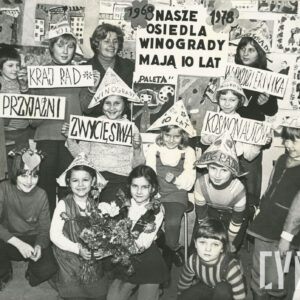 grupa dzieci z opiekunką w pomieszczeniu pełnym prac plastycznych; część dzieci nosi papierowe czapki; w tle na ścianach wiszą liczne rysunki i prace plastyczne