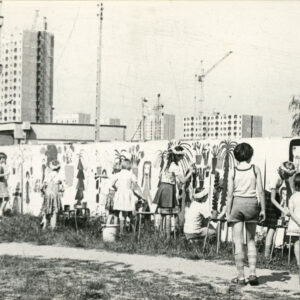 grupa dzieci malujących po płocie; w oddali widać żurawie budowalne i nowo powstające bloki z wielkiej płyty