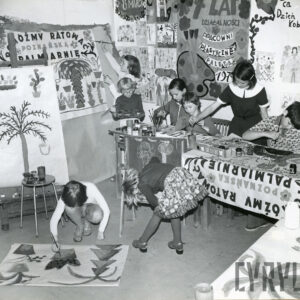 grupa dzieci malujących przy stołach i na podłodze pomieszczenia, na ścianach którego wiszą różne prace plastyczne