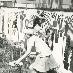 grupa dzieci malujących po płocie; w oddali widać żurawie budowalne i nowo powstające bloki z wielkiej płyty