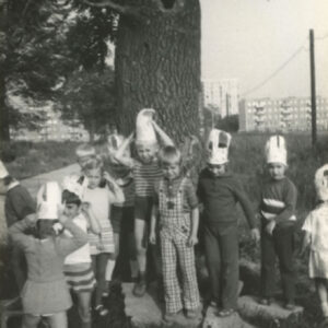 grupa przebranych dzieci, część z nich z papierowymi czapkami na głowach, na tle pnia drzewa, w głębi fotografii bloki mieszkalne