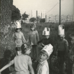 grupa przebranych dzieci, część z nich z papierowymi czapkami na głowach, bawiąca się wokół drzewa, w głębi fotografii bloki mieszkalne