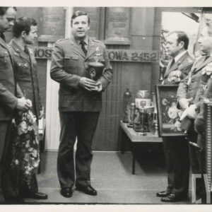 kilku mężczyzn w mundurach wojskowych stoi w pomieszczeniu, dwóch z nich, z prawej trzyma obrazy, jeden, z prawej kwiaty