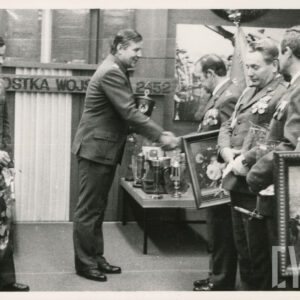 kilku mężczyzn w mundurach wojskowych, dwóch, z lewej trzyma obrazy, mężczyzna z prawej trzyma kwiaty, pośrodku dwóch mężczyzn podaje sobie ręce