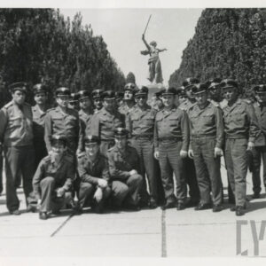 grupa mężczyzn w mundurach wojskowych stoi w alei obsadzonej drzewami, w tle pomnik kobiety z uniesionym mieczem