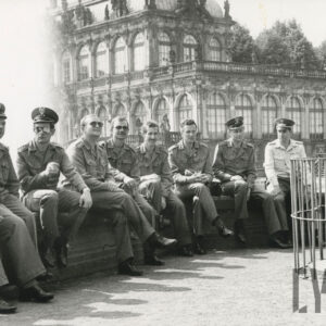 grupa mężczyzn w mundurach siedzi na murku, za nimi fontanna i bogato zdobiony budynek