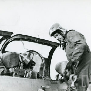 jeden mężczyzna w hełmie lotniczym siedzi w samolocie, drugi w kombinezonie lotniczym i hełmie wsiada do samolotu