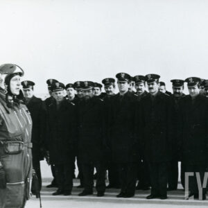 duża grupa mężczyzn w mundurach wojskowych stoi na baczność, na pierwszym planie mężczyzna w kombinezonie lotniczym i hełmie składa raport