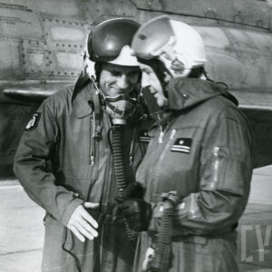 dwóch mężczyzn w kombinezonach lotniczych i hełmach, za nimi fragment samolotu wojskowego