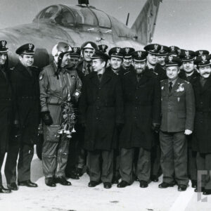 grupa mężczyzn w mundurach, płaszczach i czapkach wojskowych na tle samolotu, wśród nich pilot w hełmie i skafandrze lotniczym