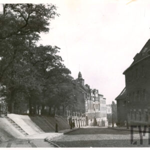 brukowana ulica, w głębi i z prawej kilkukondygnacyjne budynki, z lewej skarpa, na niej drzewa