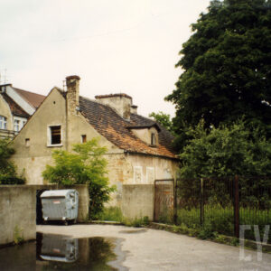 budynek z odpadającym tynkiem, skośnym dachem i kominem, na pierwszym planie płot częściowo murowany, częściowo metalowy, przy płocie kontener na śmieci