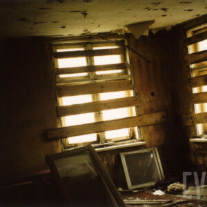 pomieszczenie z oknami, zabitymi deskami, na podłodze porozrzucany gruz i fragment ram okiennych, na suficie odpadająca farba