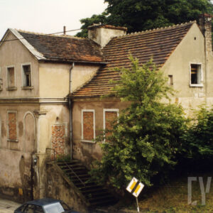 budynek z zamurowanymi oknami i wejściem, na ścianach odpadający tynk, przy budynku wysokie krzaki