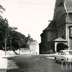 asfaltowa ulica, z prawej samochód osobowy, z prawej fragment budynku szkoły, w głębi kilkukondygnacyjne kamienice