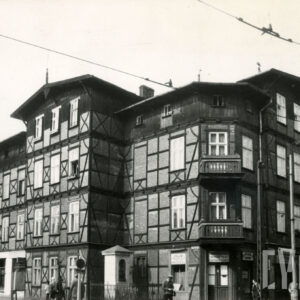 czterokondygnacyjny, drewniano-ceglany budynek z drewnianym poddaszem, w narożniku dwie kondygnacje z drewnianymi balkonami, w wyciętym narożniku budynku kapliczka ogrodzona niskim płotem, chodnikiem spacerują ludzie