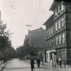 brukowana ulica, obsadzona drzewami, z prawej czterokondygnacyjny, drewniano-ceglany budynek, w wyciętym narożniku budynku kapliczka ogrodzona niskim płotem, ulicą i chodnikiem spacerują ludzie