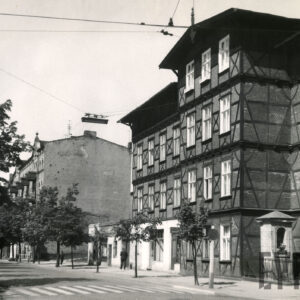 brukowana ulica, obsadzona drzewami, z prawej czterokondygnacyjny, drewniano-ceglany budynek, w wyciętym narożniku budynku kapliczka ogrodzona niskim płotem