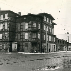 dwupiętrowy drewniano-ceglany dom narożny z mieszkalnym poddaszem, na pierwszym planie brukowana ulica z torami tramwajowymi, resztki zalegającego śniegu