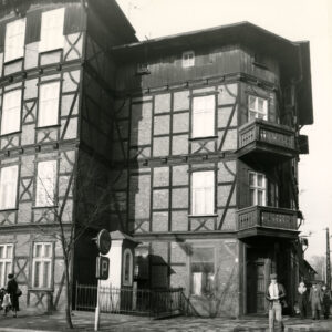 fragment czterokondygnacyjnego, drewniano-ceglanego budynku z drewnianym poddaszem, w narożniku dwie kondygnacje z drewnianymi balkonami, w wyciętym narożniku budynku kapliczka ogrodzona niskim płotem, chodnikiem spacerują ludzie