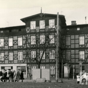 czterokondygnacyjny, drewniano-ceglany budynek z drewnianym poddaszem, w narożniku dwie kondygnacje z drewnianymi balkonami, z lewej ludzie stoją w kolejce