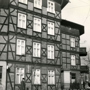 fragment czterokondygnacyjnego, drewniano-ceglanego budynku z drewnianym poddaszem, w narożniku, z prawej dwie kondygnacje z drewnianymi balkonami, w wyciętym narożniku budynku kapliczka ogrodzona niskim płotem