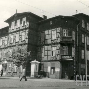 czterokondygnacyjny, drewniano-ceglany budynek z drewnianym poddaszem, w narożniku dwie kondygnacje z drewnianymi balkonami, w wyciętym narożniku budynku kapliczka ogrodzona niskim płotem, chodnikiem spacerują ludzie