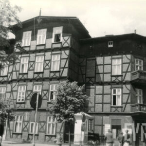 czterokondygnacyjny, drewniano-ceglany budynek z drewnianym poddaszem, w narożniku dwie kondygnacje z drewnianymi balkonami, w wyciętym narożniku budynku kapliczka ogrodzona niskim płotem, chodnikiem spacerują ludzie