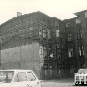 czterokondygnacyjny, drewniano-ceglany budynek, na placu przed budynkiem zaparkowane samochody