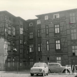 czterokondygnacyjny, drewniano-ceglany budynek, na placu przed budynkiem zaparkowane samochody