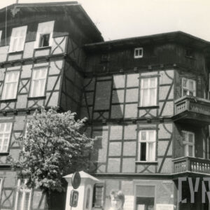 czterokondygnacyjny, drewniano-ceglany budynek z drewnianym poddaszem, w narożniku dwie kondygnacje z drewnianymi balkonami, w wyciętym narożniku budynku kapliczka