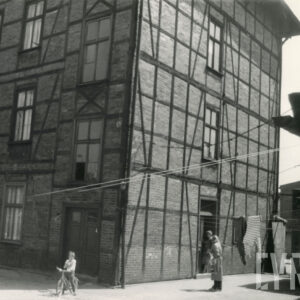 czterokondygnacyjny, drewniano-ceglany budynek, przy wejściu stoją dwie osoby, z lewej dziecko na rowerze