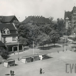 na pierwszym planie fragment placu, na nim małe kioski, za nimi ulica, w głębi budynki murowane i drzewa