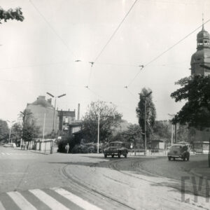 brukowana ulica z torami tramwajowymi, jadą dwa samochody osobowe, w głębi skupisko drzew, z prawej wieża kościoła, w głębi, nieco z lewej kamienice