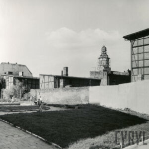 na pierwszym planie mur, za nim fragmenty drewniano-ceglanych budynków, w głębi wieża kościoła, wokół niej rusztowania