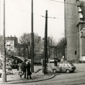 na pierwszym planie brukowana ulica z torami tramwajowymi, z lewej stragany, spacerujący ludzi, z prawej fragment kościoła