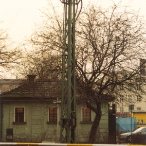 niski budynek z trzema oknami, przy nim drzewo, na pierwszym planie kratownicowy słup z trakcją tramwajową i metalowa barierka odgradzająca chodnik od ulicy